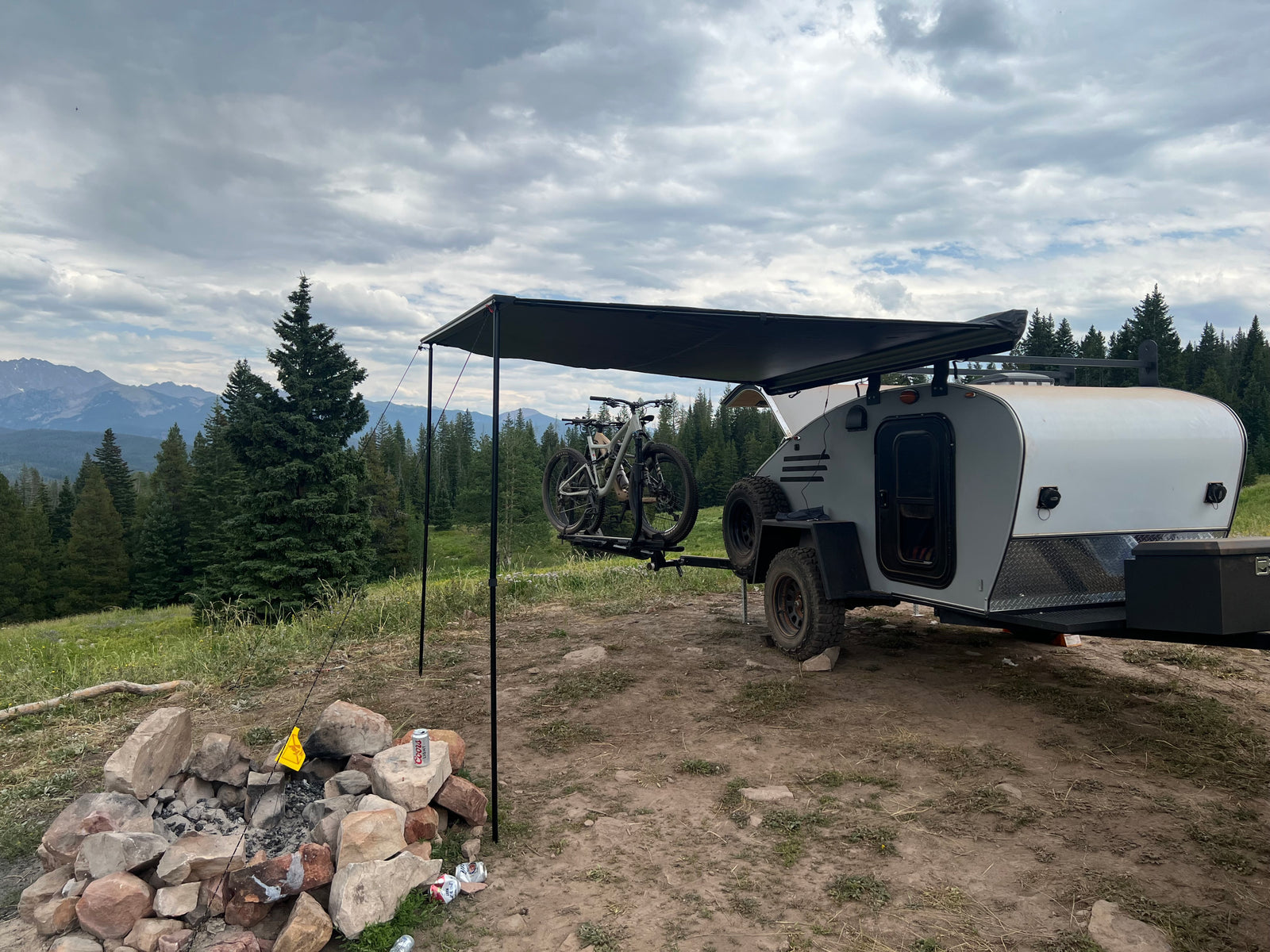 Scenic RV rental campsite: A teardrop trailer with bikes, an awning, and a fire pit, surrounded by pine trees and mountains under cloudy skies.