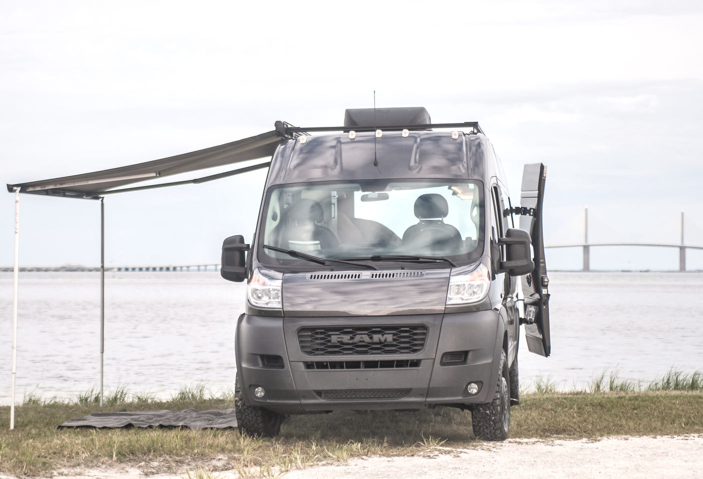 Explore with this dark grey Ram ProMaster camper van. Awning deployed by water with bridge view. Ideal rv rental for adventure!