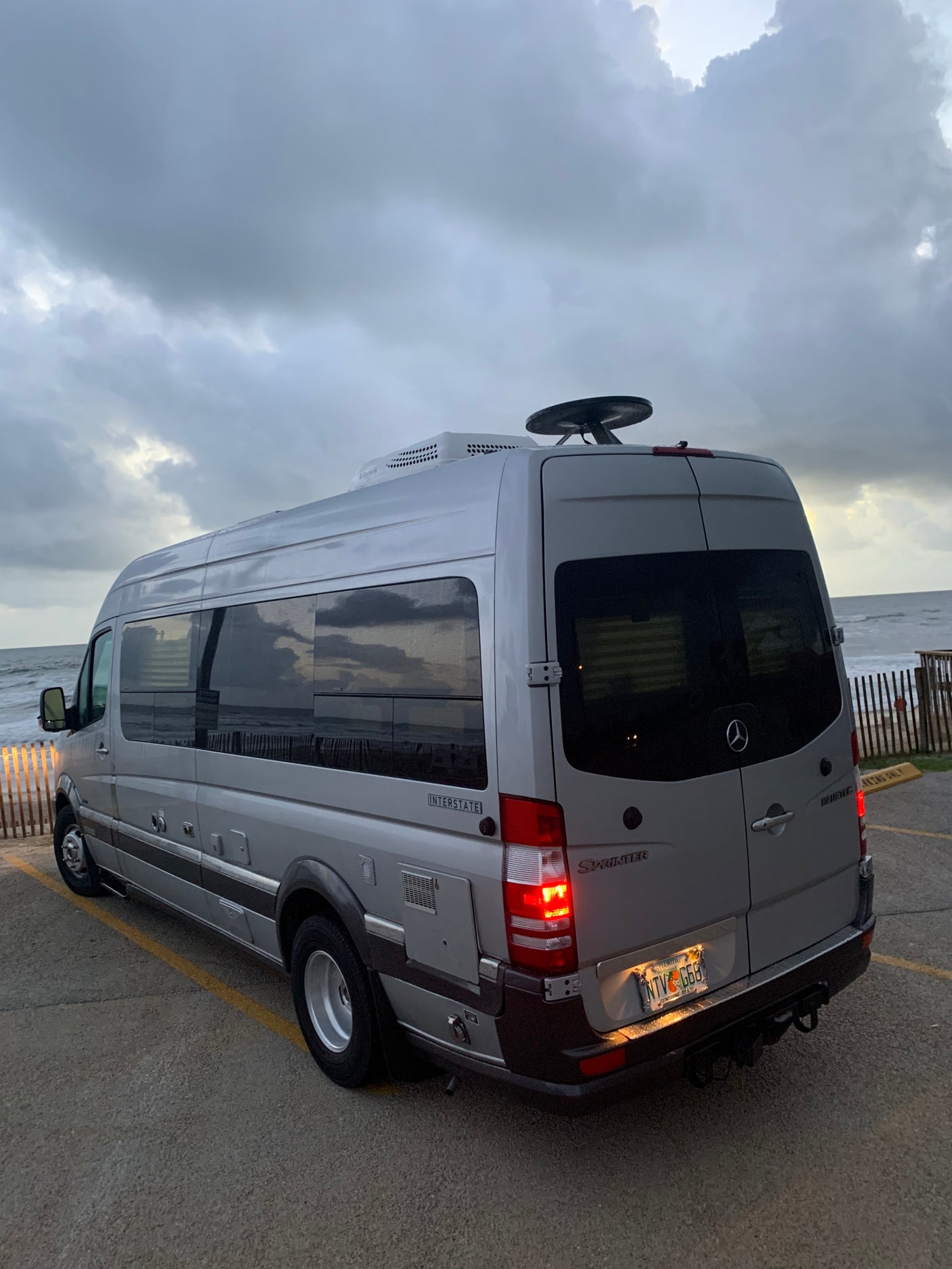 An RV rental, a silver Mercedes-Benz Sprinter van, parked by the overcast beach. Perfect for coastal adventures!