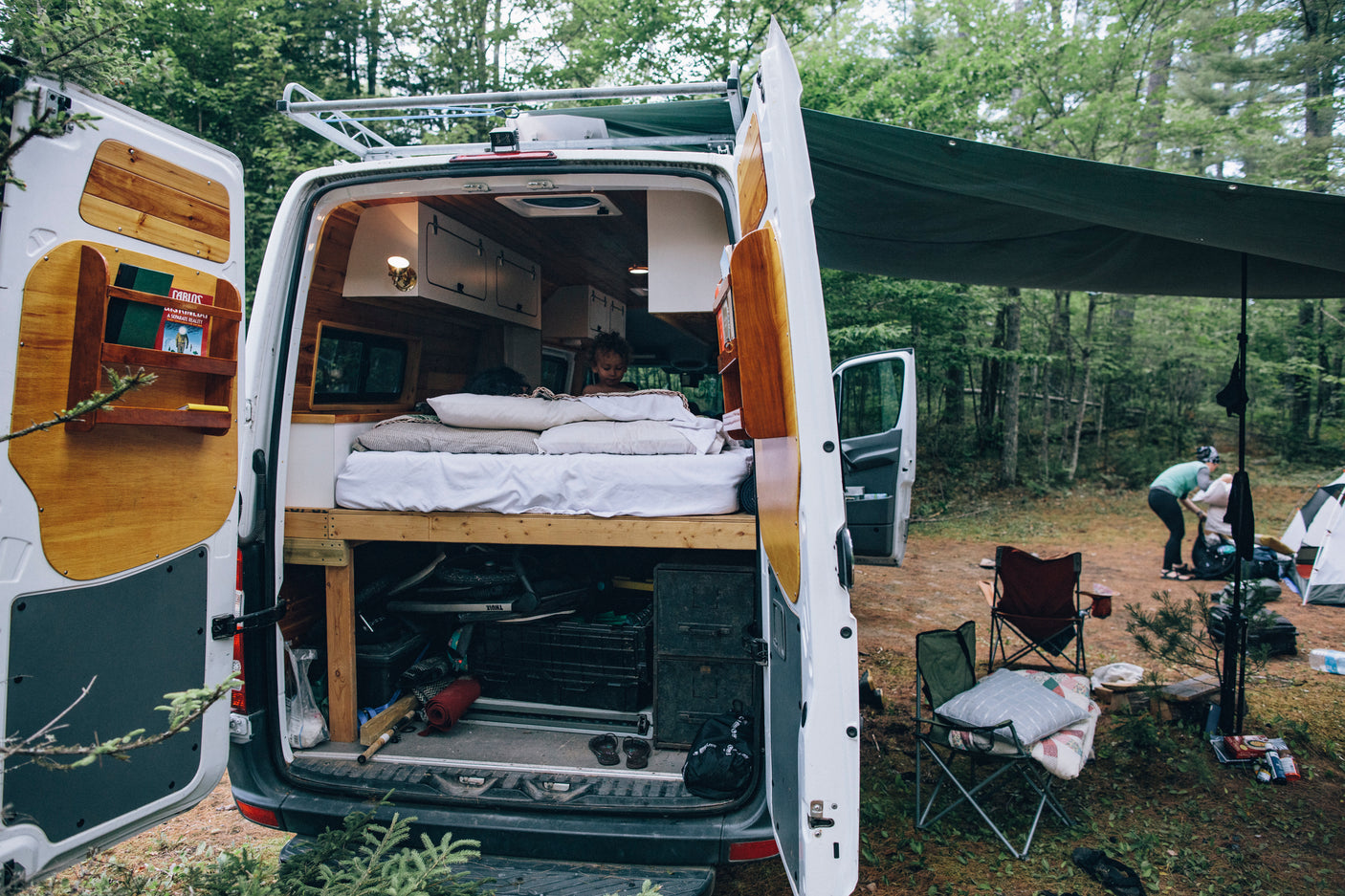 Experience forest camping with an RV rental! A child is cozy inside the custom camper van, while an adult sets up camp outside.