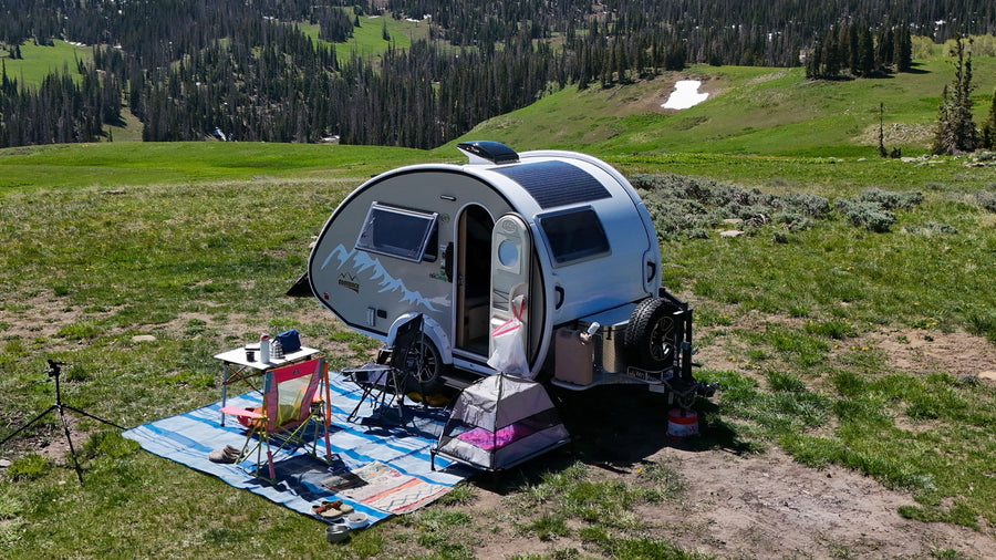 Teardrop RV rental set up for off-grid camping in a scenic mountain meadow with pine trees and snow patches.