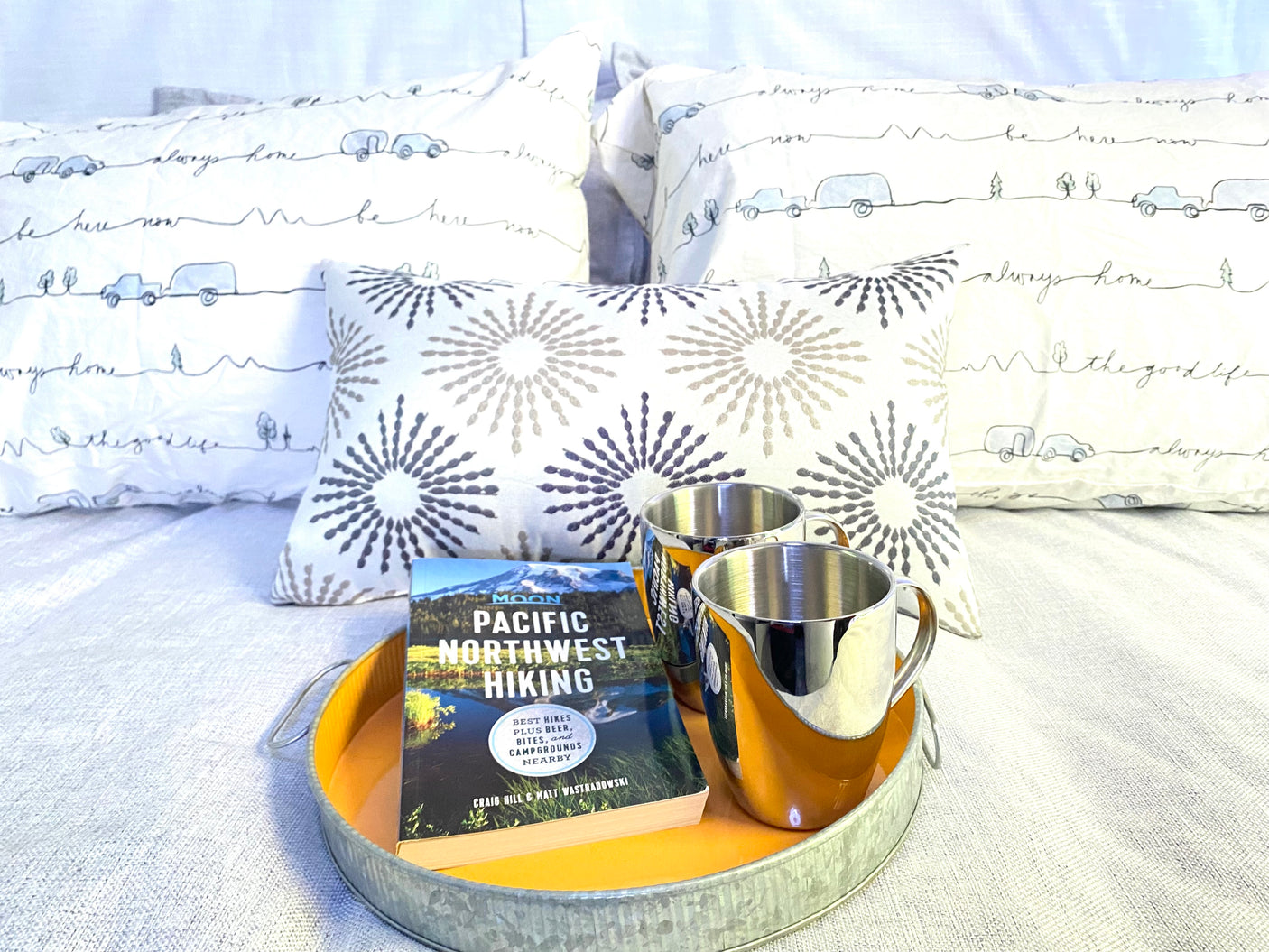 Cozy bed with travel-themed pillows, a 'Pacific Northwest Hiking' guide, & mugs on a tray for your RV rental adventure.