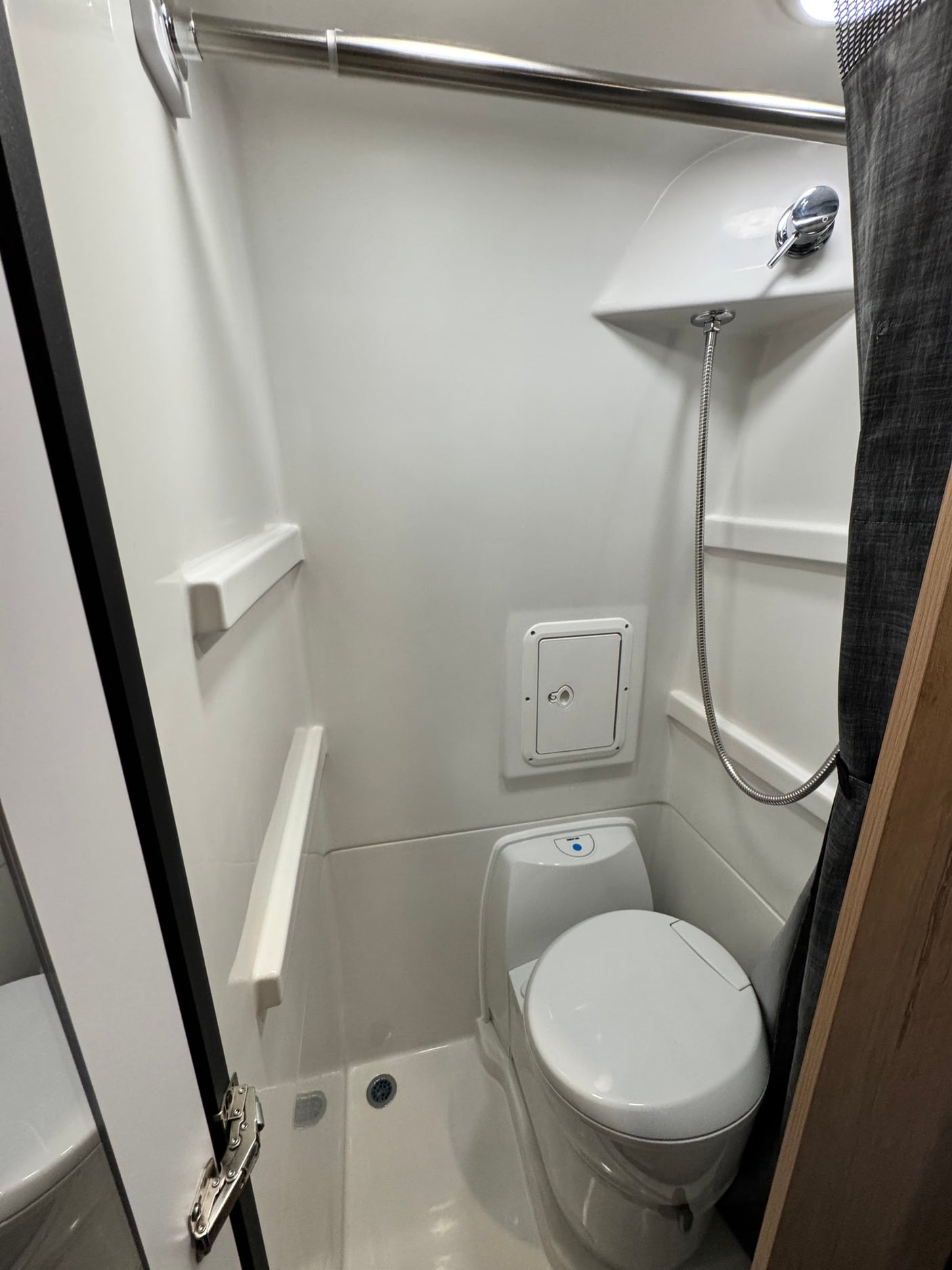 This RV rental wet bath features a compact toilet, shower with flexible hose, built-in shelves, and drain on a white interior.