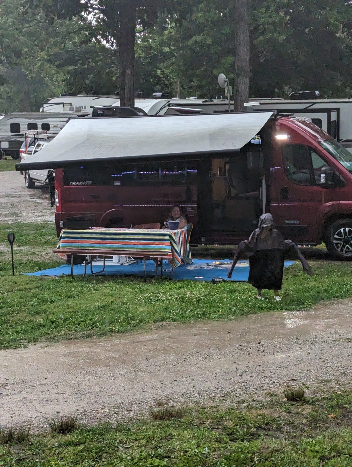 Rainy campground scene: a red RV rental with an awning provides shelter. A person is seated at a striped table, another in a poncho.