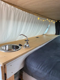 RV rental interior: Compact camper van with a wooden counter, small sink, faucet, and string lights over pleated fabric.