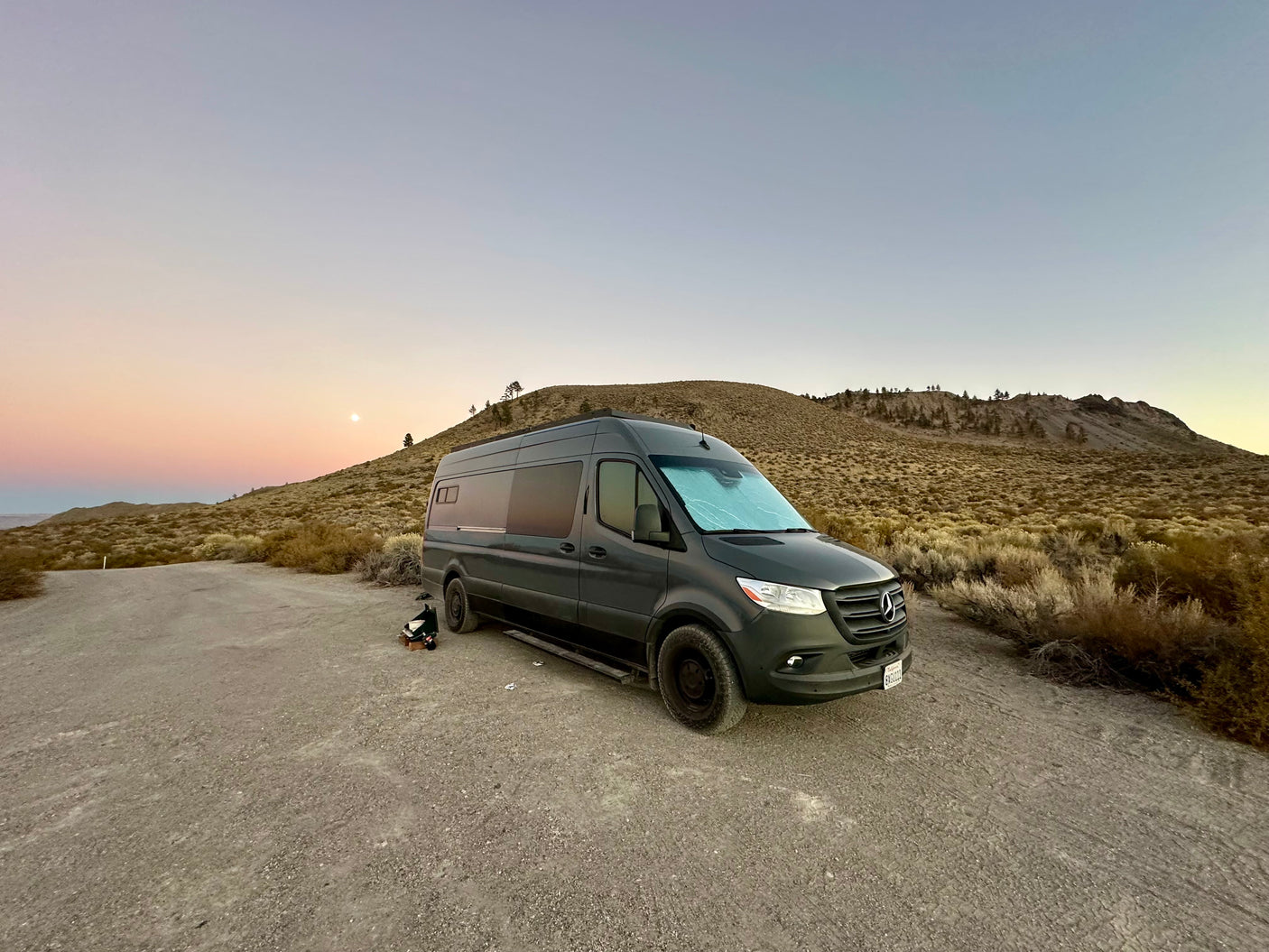 Beautiful sunset over an rv rental: a dark grey Sprinter van parked on a dirt road in a desert landscape with hills and moon.