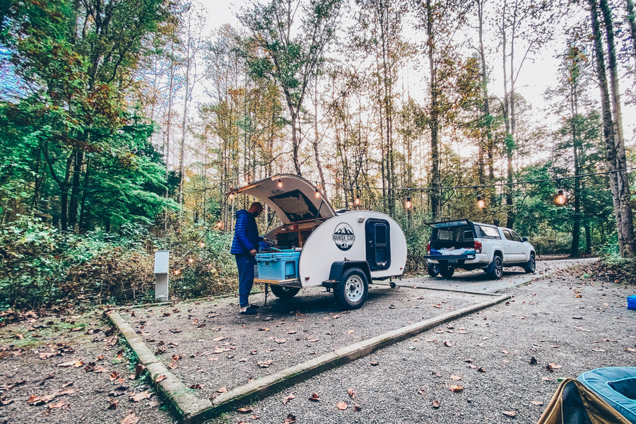 Cozy RV rental campsite in the forest: A person cooks at a teardrop trailer, with a truck & string lights nearby.
