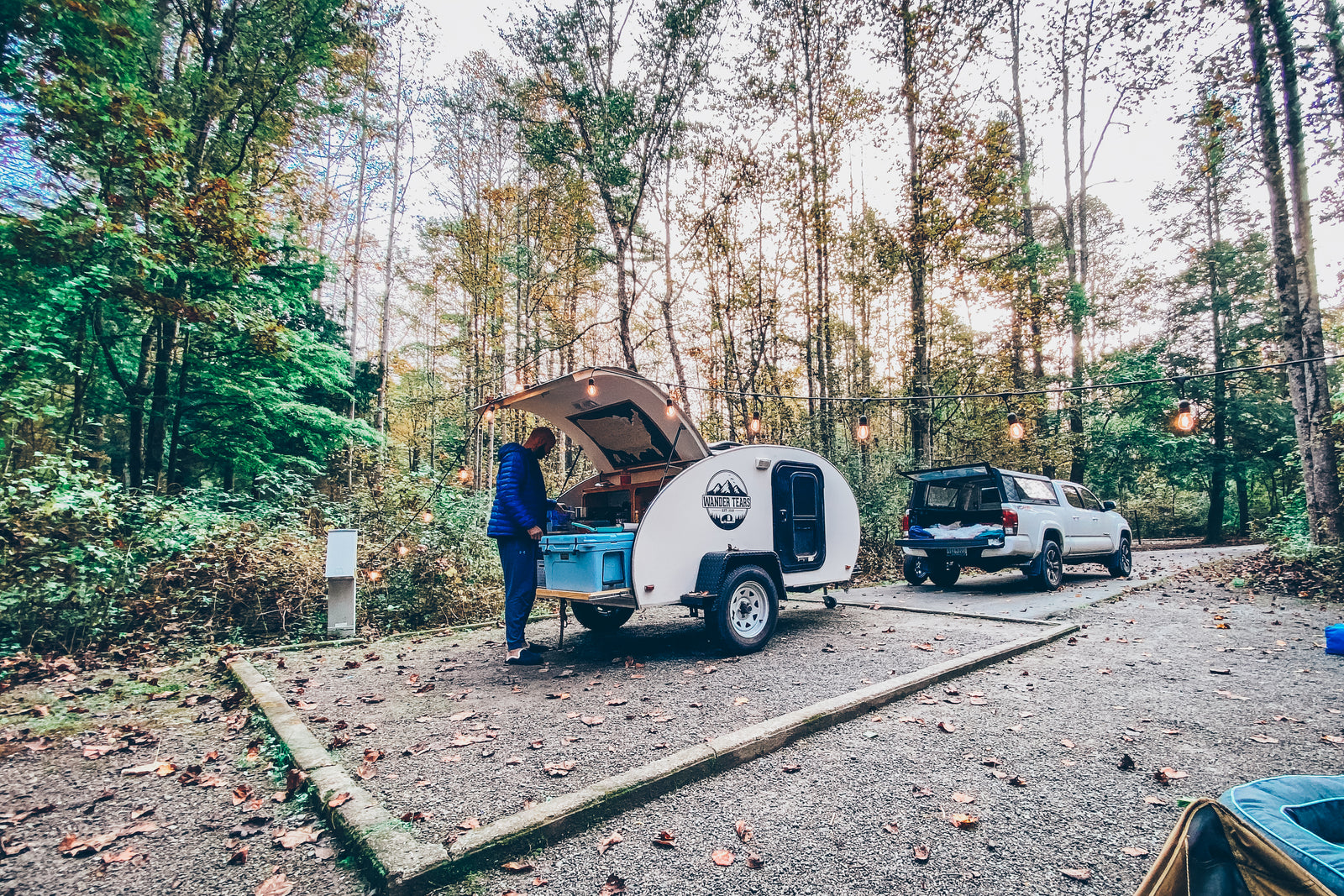 Cozy RV rental campsite in the forest: A person cooks at a teardrop trailer, with a truck & string lights nearby.
