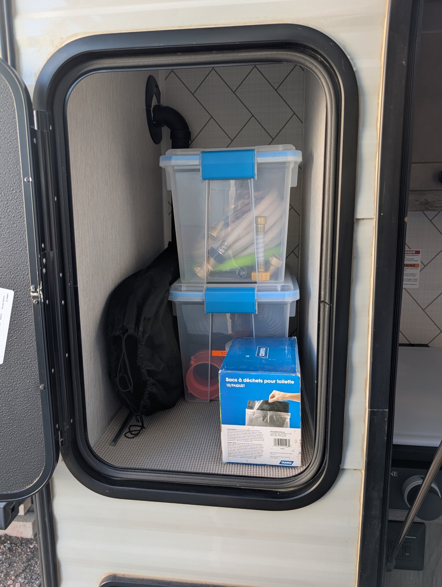 An RV rental's organized exterior storage, revealing clear bins with hoses, fittings, and a box of toilet waste bags.