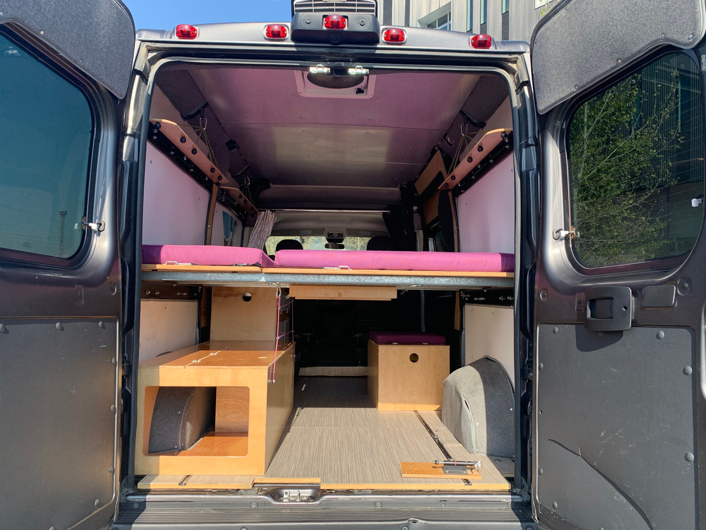 RV rental: Interior of a camper van with open back doors, showcasing a raised bed with purple cushions and wood storage.