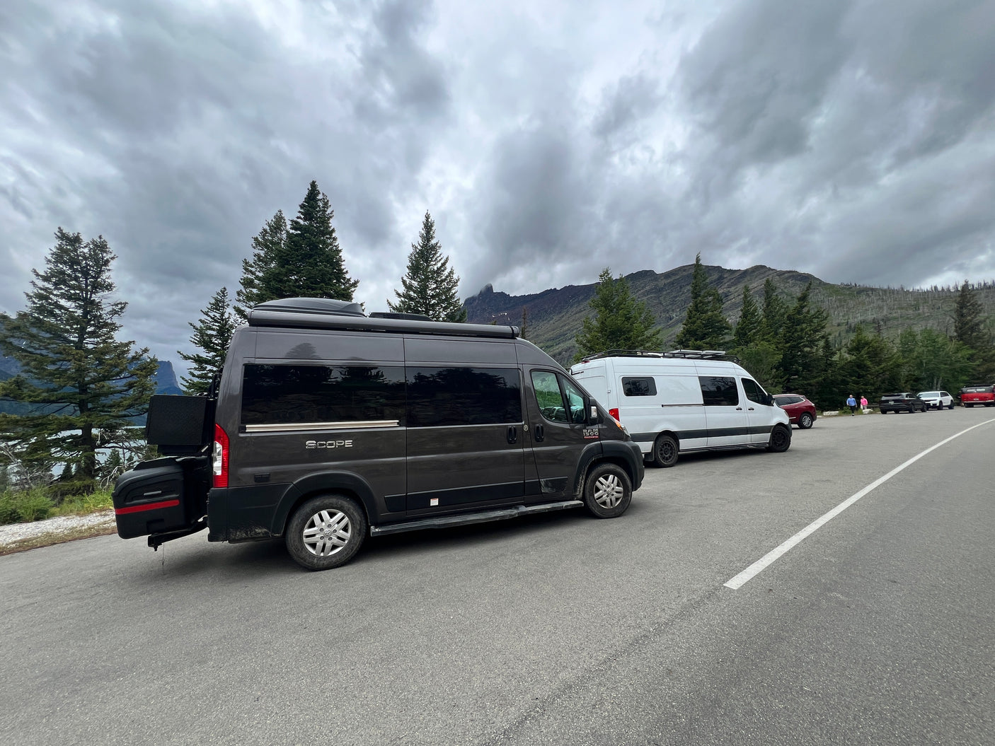 Dark grey and white RV rental vans parked by a scenic mountain lake under cloudy skies.