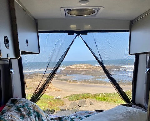 Inside an RV rental: a screened view of a sandy beach, ocean, and rocks, with a white dog on the shore. Ideal for travel.