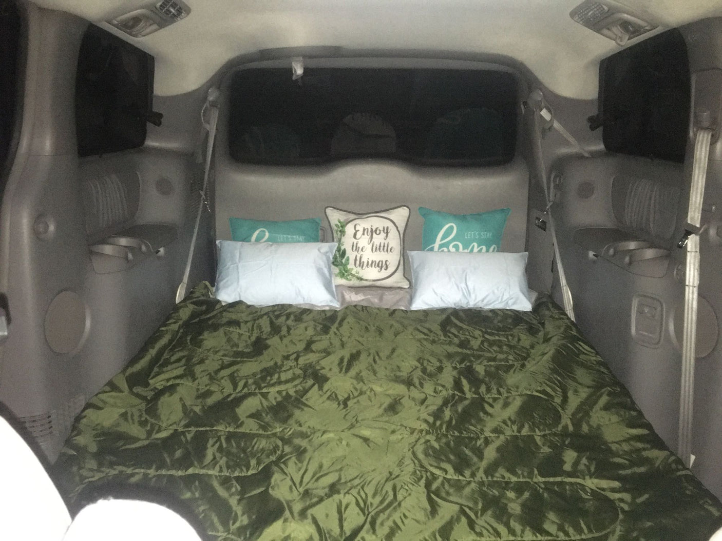 Cozy vehicle bed setup in a gray minivan/SUV, with a green sleeping bag and decorative pillows. Great for car camping or an RV rental experience.