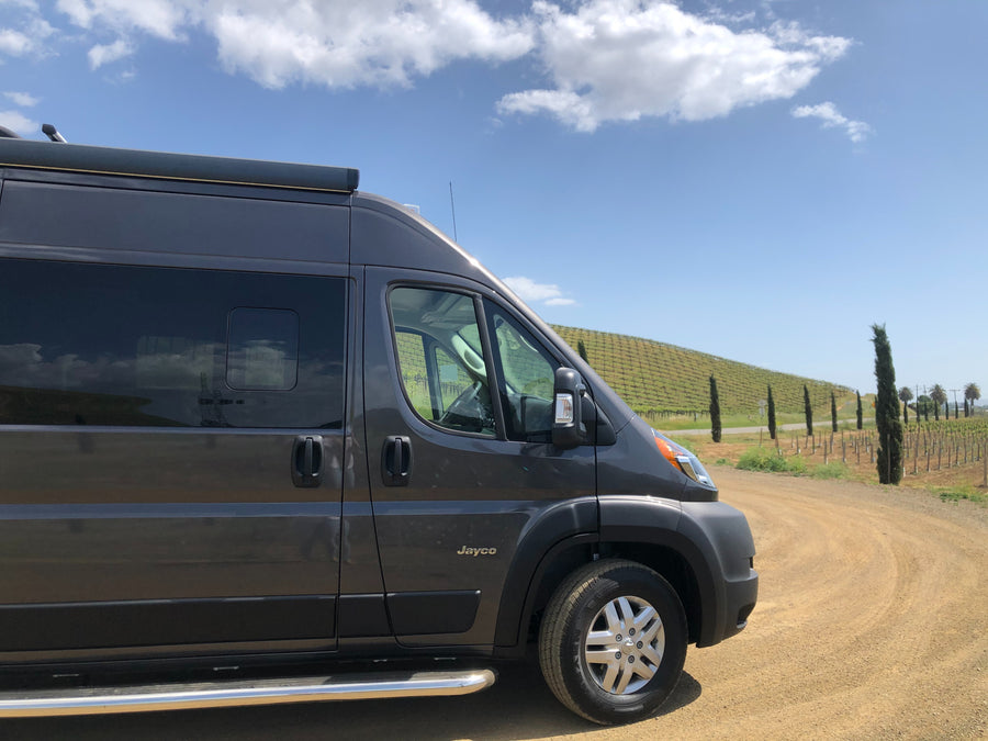 A dark gray Jayco RV rental camper van on a dirt road, next to a sunny vineyard with cypress trees. Perfect for a rural trip.