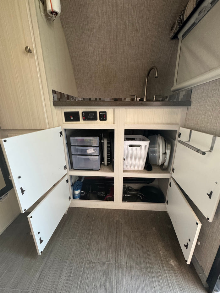 Inside an RV rental: a compact kitchen area with sink, counter, and open organized cabinets revealing storage solutions.