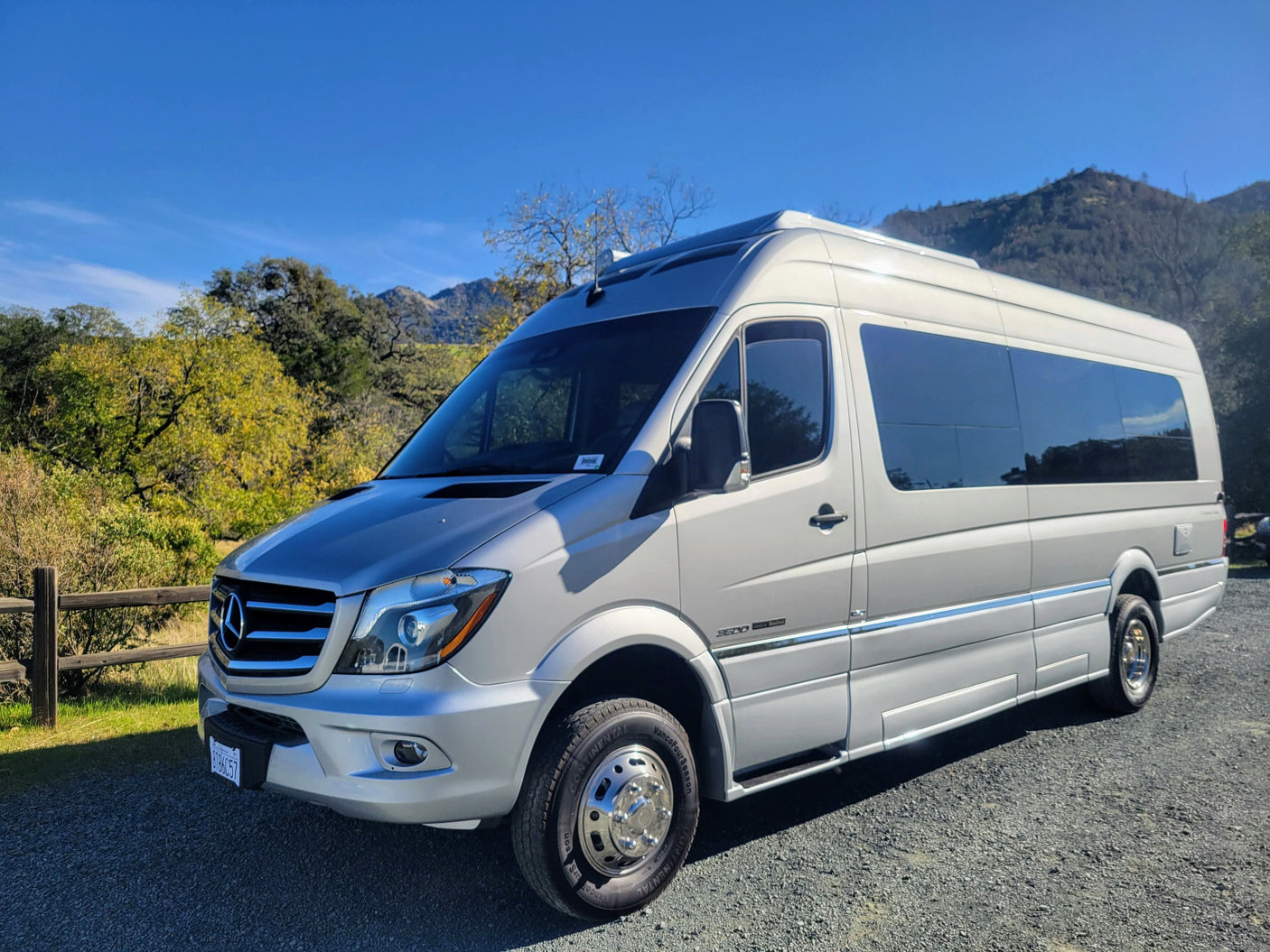 Light-colored Mercedes-Benz Sprinter RV rental parked on gravel, with green trees & distant mountains under a blue sky.
