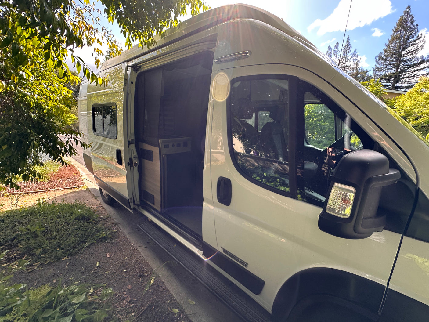 White Winnebago rv rental camper van with open side door showing interior, parked amidst trees on a sunny day.
