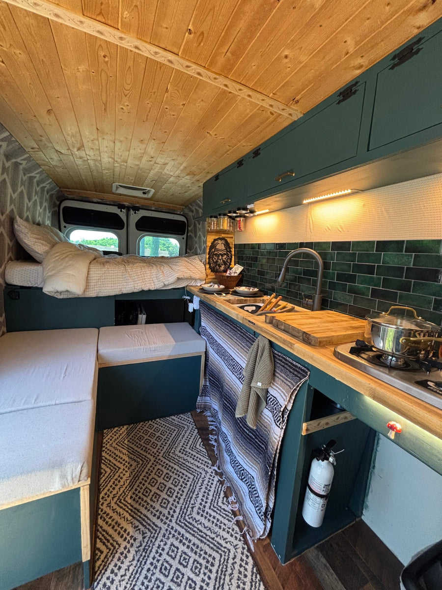Cozy converted van interior for RV rental: bed, kitchen with green tiles, wood ceiling, and patterned rug.