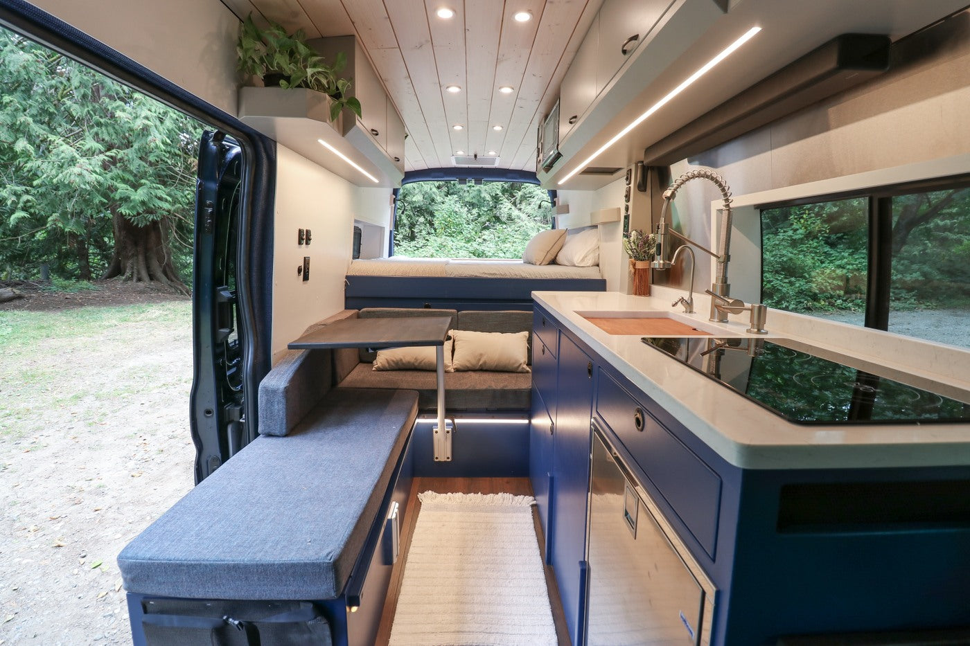 Modern rv rental camper van interior featuring kitchen, seating, bed, and open side door to lush forest.