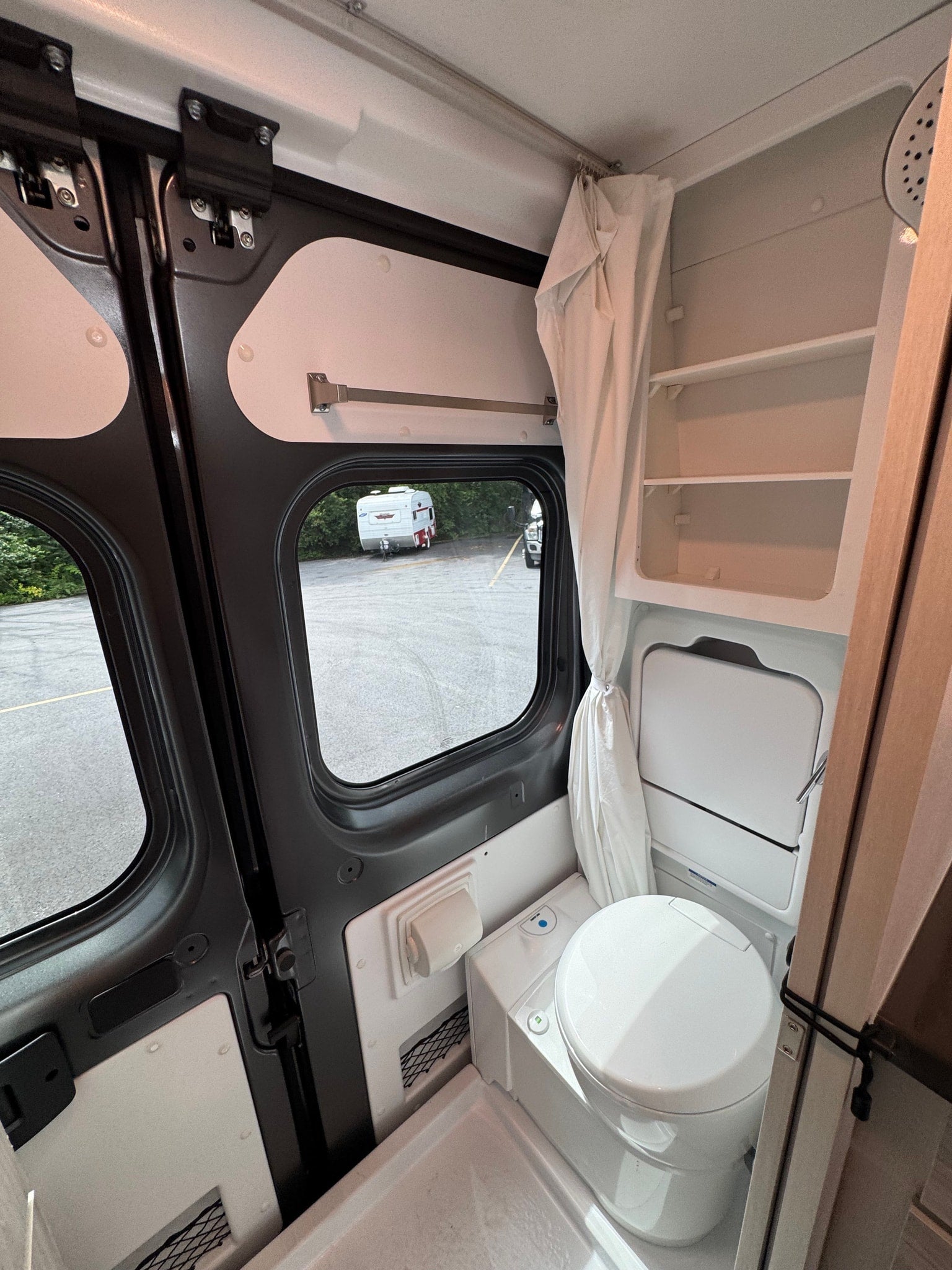 Interior of an RV rental camper van, showing the compact bathroom with a toilet, shower pan, and shelves. Parked outside with a trailer.