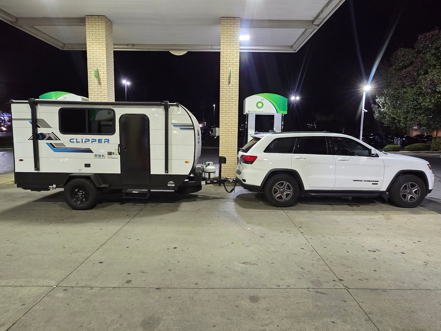 RV rental adventure: White Jeep Grand Cherokee towing a Clipper travel trailer at a brightly lit BP gas station at night.