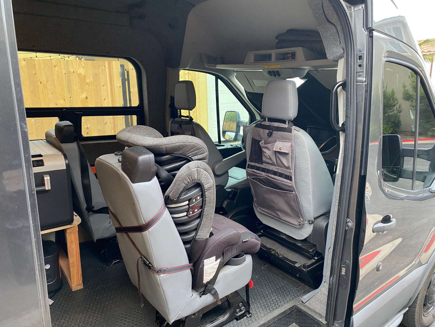Inside a gray RV rental van, highlighting a child car seat, portable fridge on a stand, and seat-back organizer.