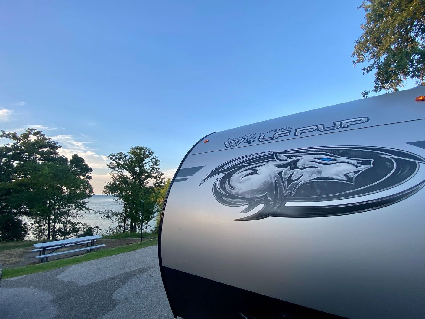 Lakeside bliss with our RV rental! The front of a 'Wolf Pup' travel trailer, parked by water, trees & a picnic table.