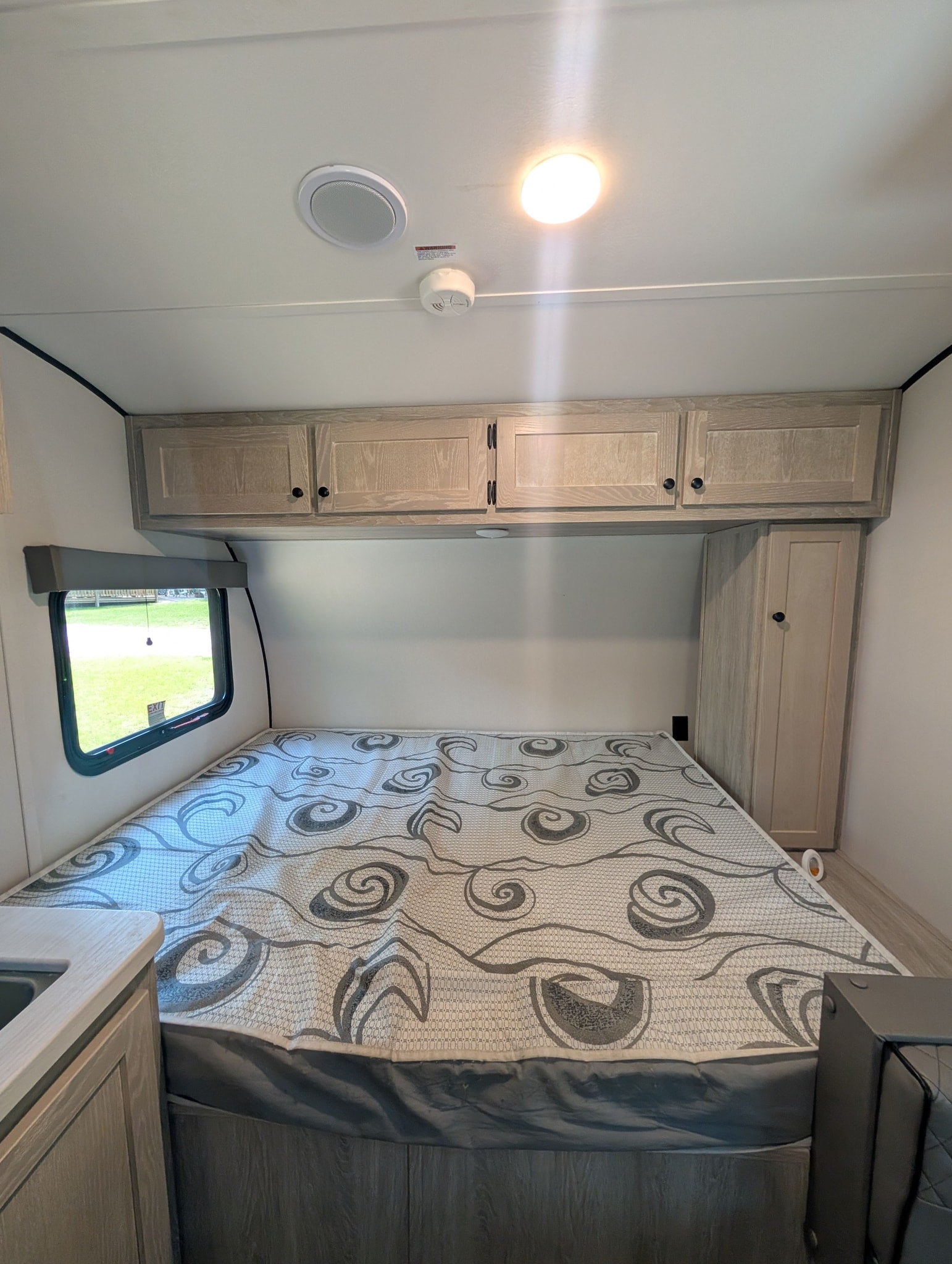 Inside an RV rental: a neat bedroom with a patterned mattress, wooden overhead cabinets, and a side window.