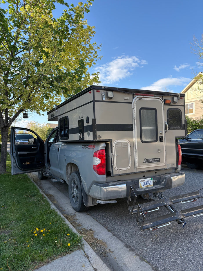 Grey pickup truck with a "Four Wheel Hawk" pop-up camper (like an RV rental) parked on a sunny street.