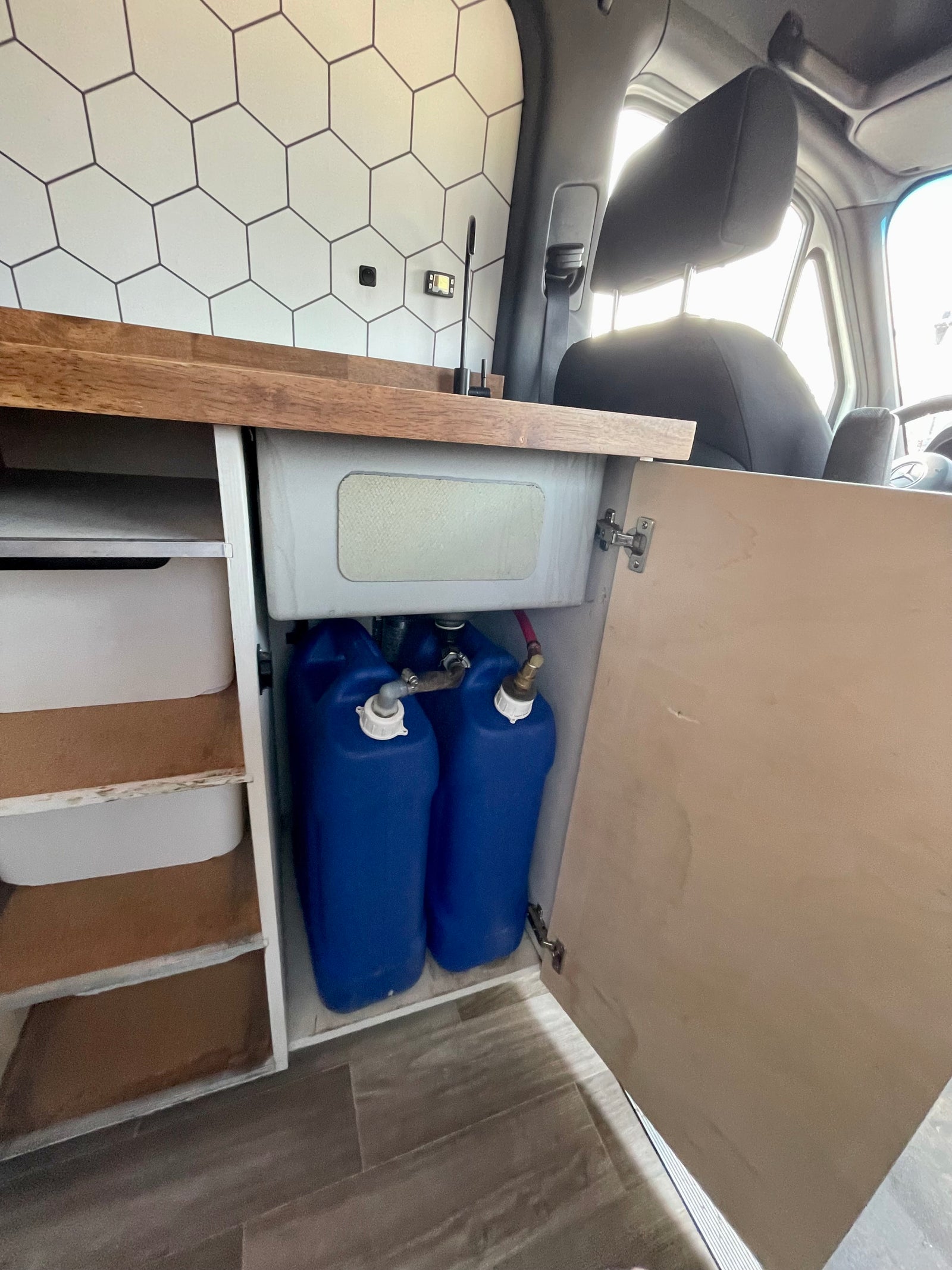 This RV rental interior shows a compact camper van kitchen with a sink, two blue water tanks, and organized storage.