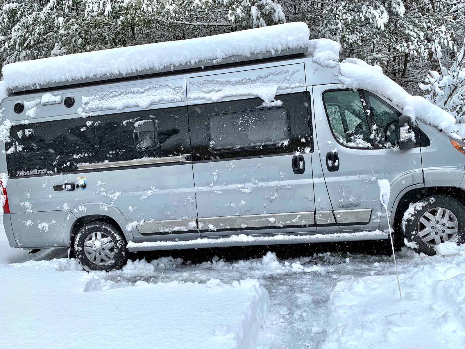 This silver Winnebago Travato rv rental is buried in fresh snow during a winter storm, with snowy trees in the background.