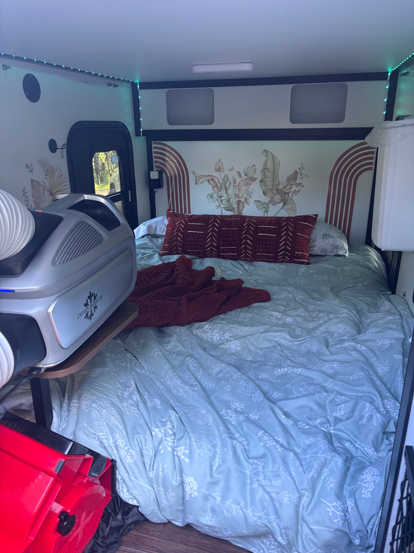 Cozy RV rental interior featuring a bed with botanical decor, LED lighting, and a portable Zero Breeze AC unit for comfort.