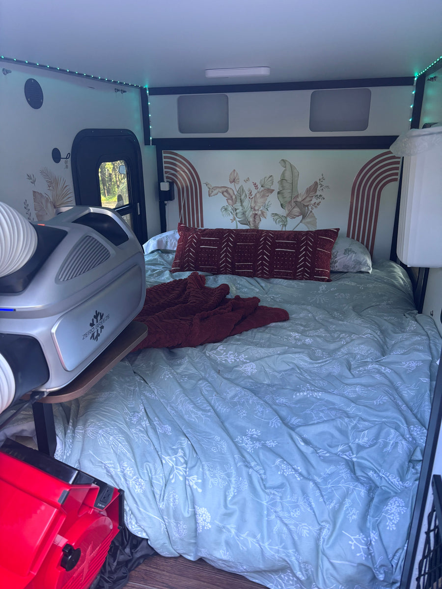 Cozy RV rental interior featuring a bed with botanical decor, LED lighting, and a portable Zero Breeze AC unit for comfort.