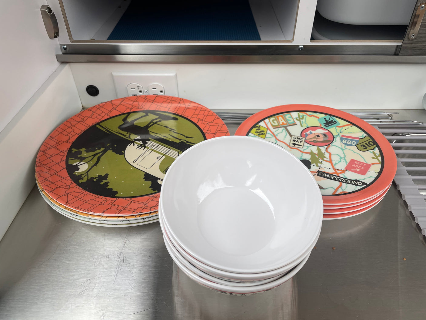 RV rental kitchen prep: Colorful camping plates & white bowls stacked on a metallic counter, ready for adventure.