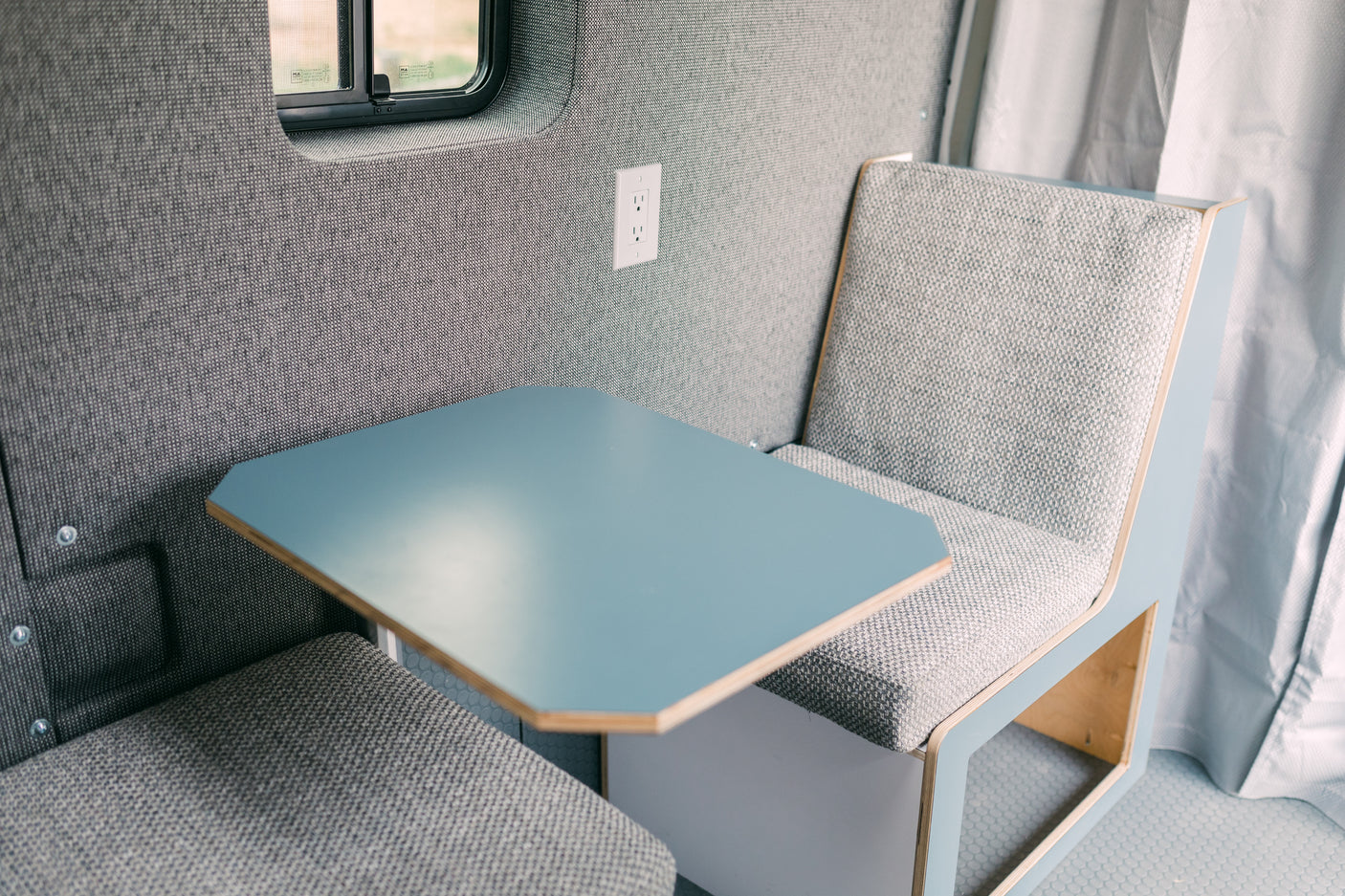Modern RV rental interior with light gray upholstered seating, blue table, and window. A compact, efficient travel space.