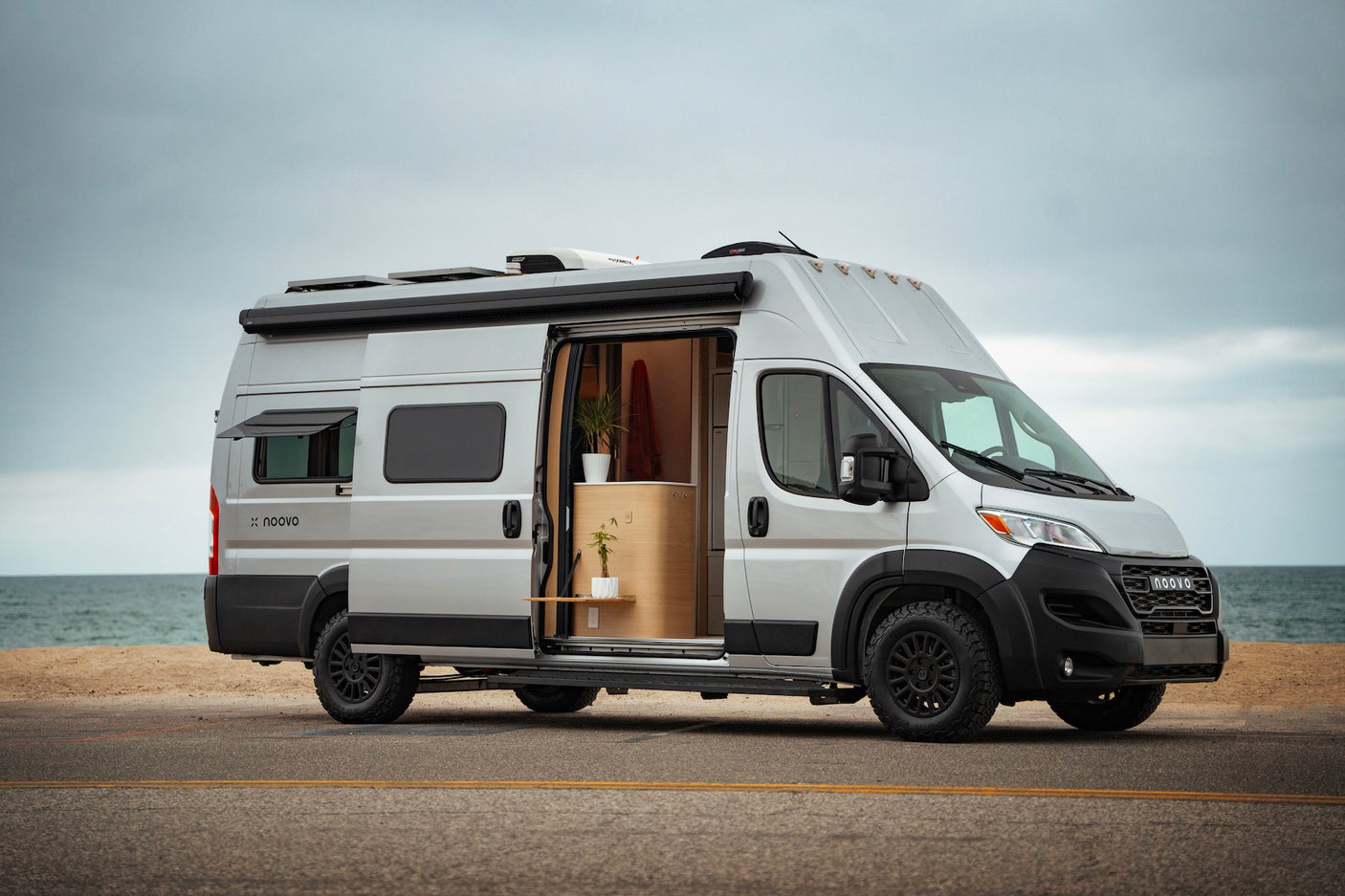 Silver Noovo camper van RV rental parked by the ocean with its door open, revealing a modern, plant-filled interior.