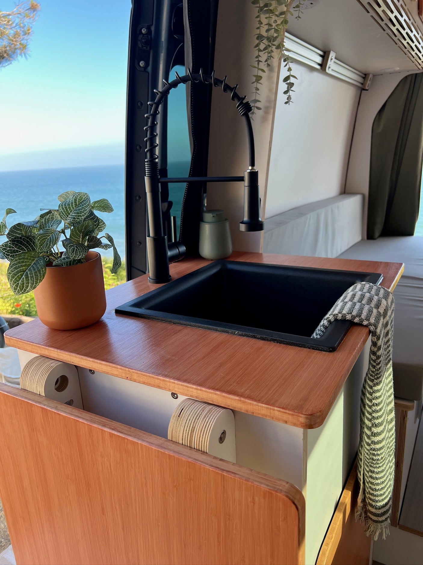 Enjoy your RV rental escape! This modern camper van kitchen boasts a black sink, wood counter, and an amazing ocean view.