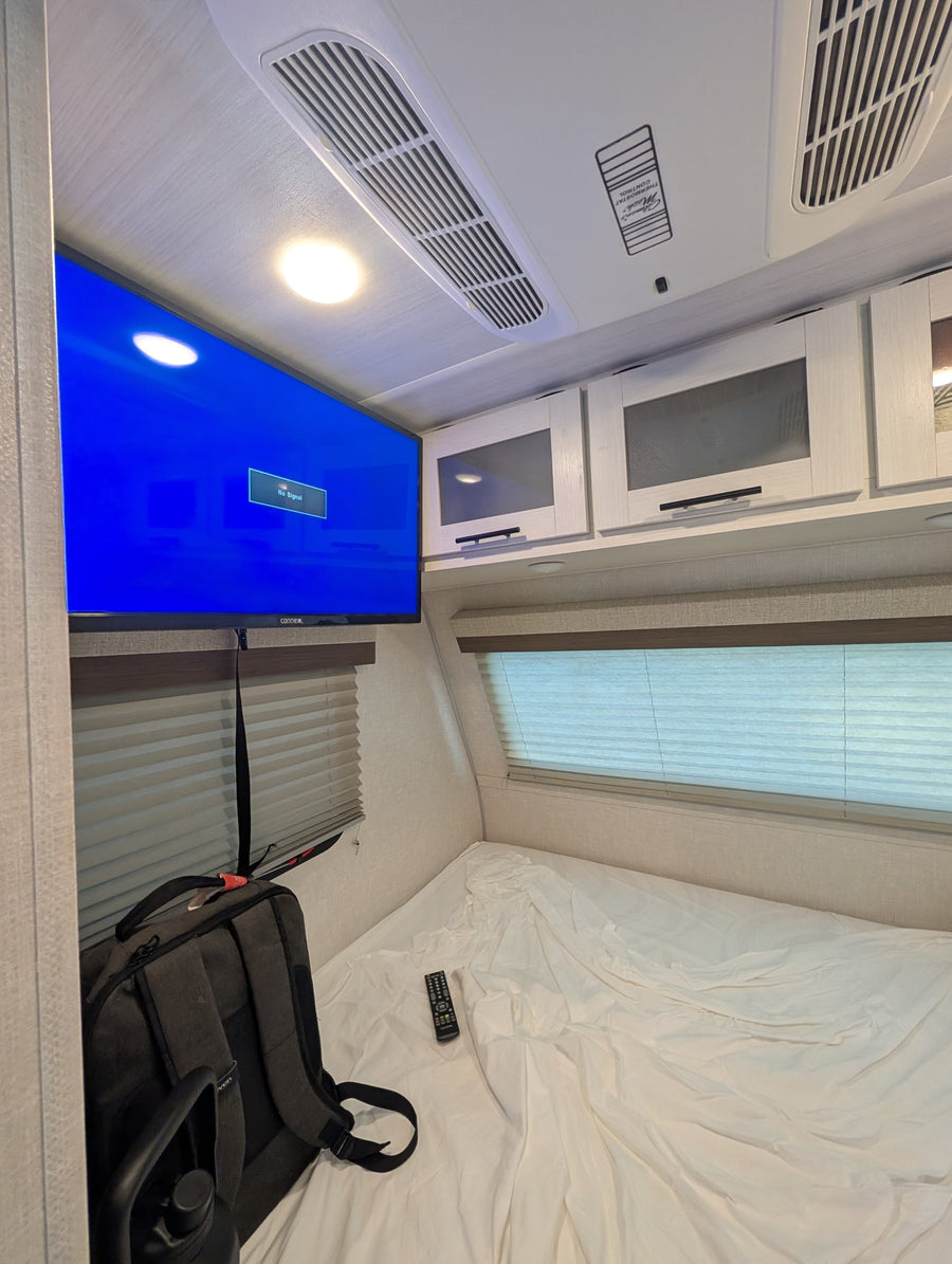 Inside an RV rental bedroom: A mounted TV, AC unit, overhead cabinets, and a bed with a remote and backpack.