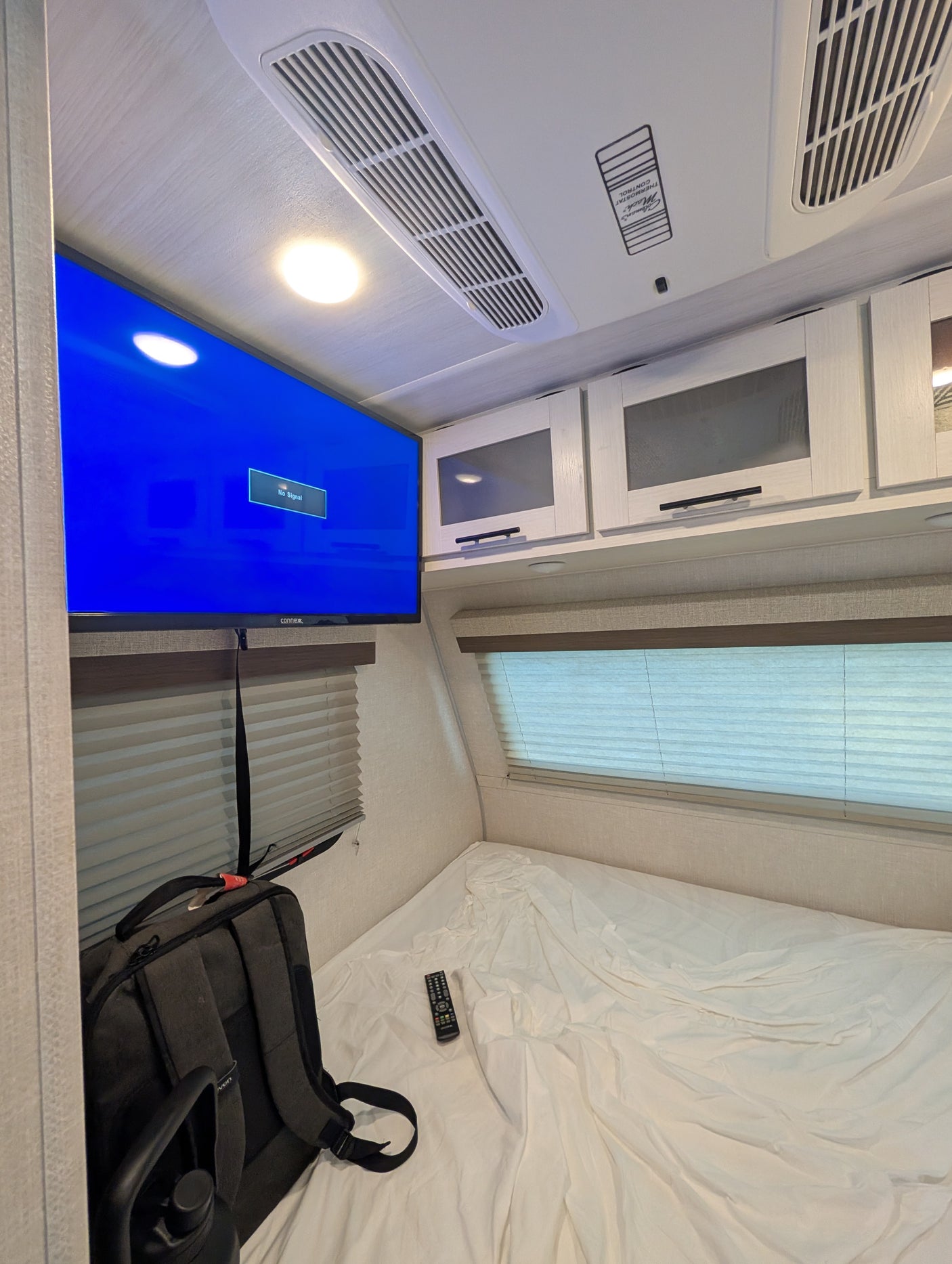 Inside an RV rental bedroom: A mounted TV, AC unit, overhead cabinets, and a bed with a remote and backpack.