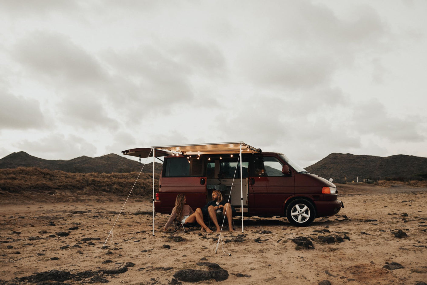 Relaxing rv rental adventure: A couple unwinds in front of their maroon camper van with lights, parked on sandy terrain near mountains.