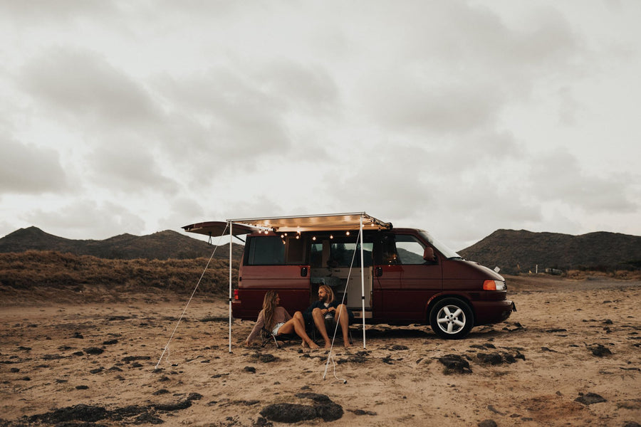 Relaxing rv rental adventure: A couple unwinds in front of their maroon camper van with lights, parked on sandy terrain near mountains.