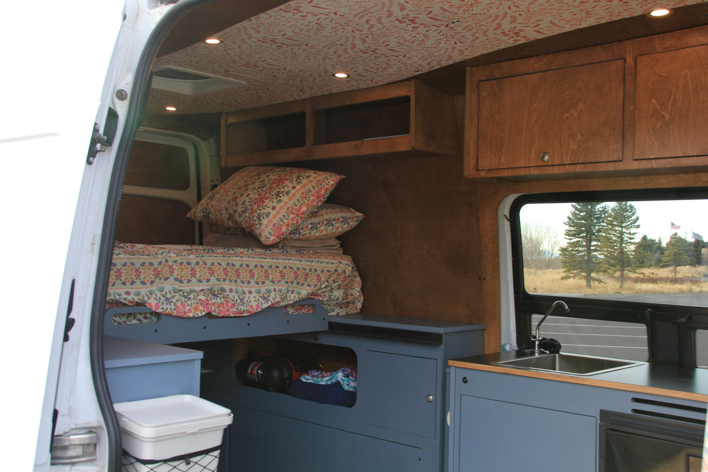 This rv rental shows a cozy, customized interior with a floral bed, blue storage, kitchen, and wood cabinets.