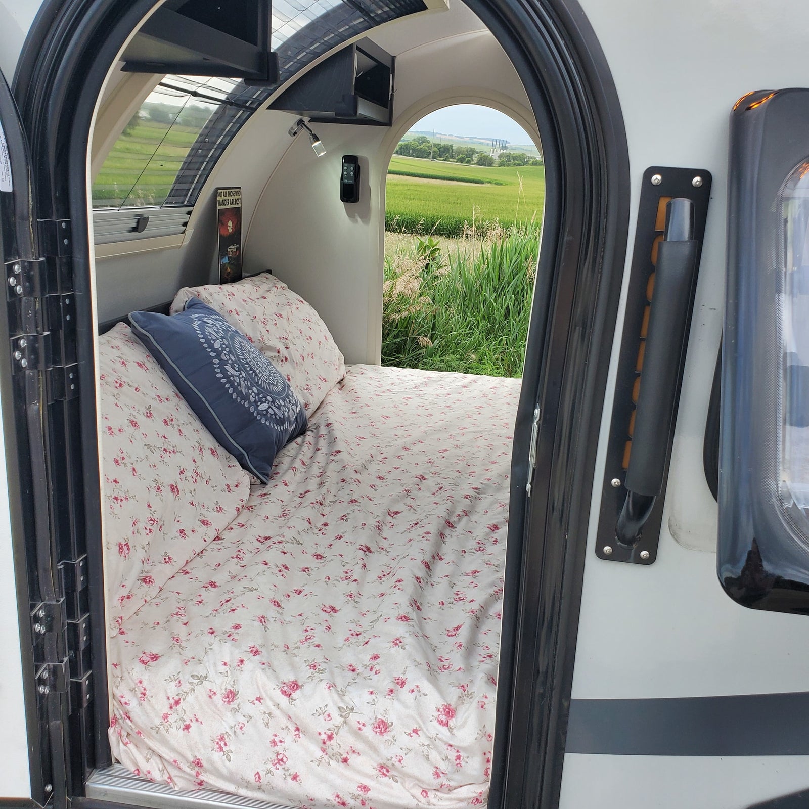 Inside a cozy RV rental: a bed with floral sheets, blue pillow, and view of green fields.