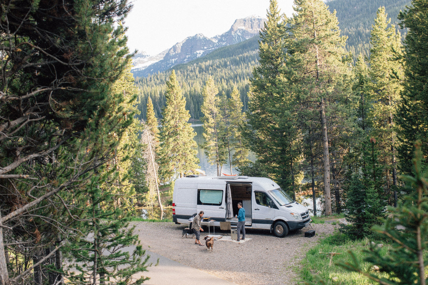 RV rental parked by a serene lake in a forest with mountains, two people and two dogs enjoying the outdoor setting.