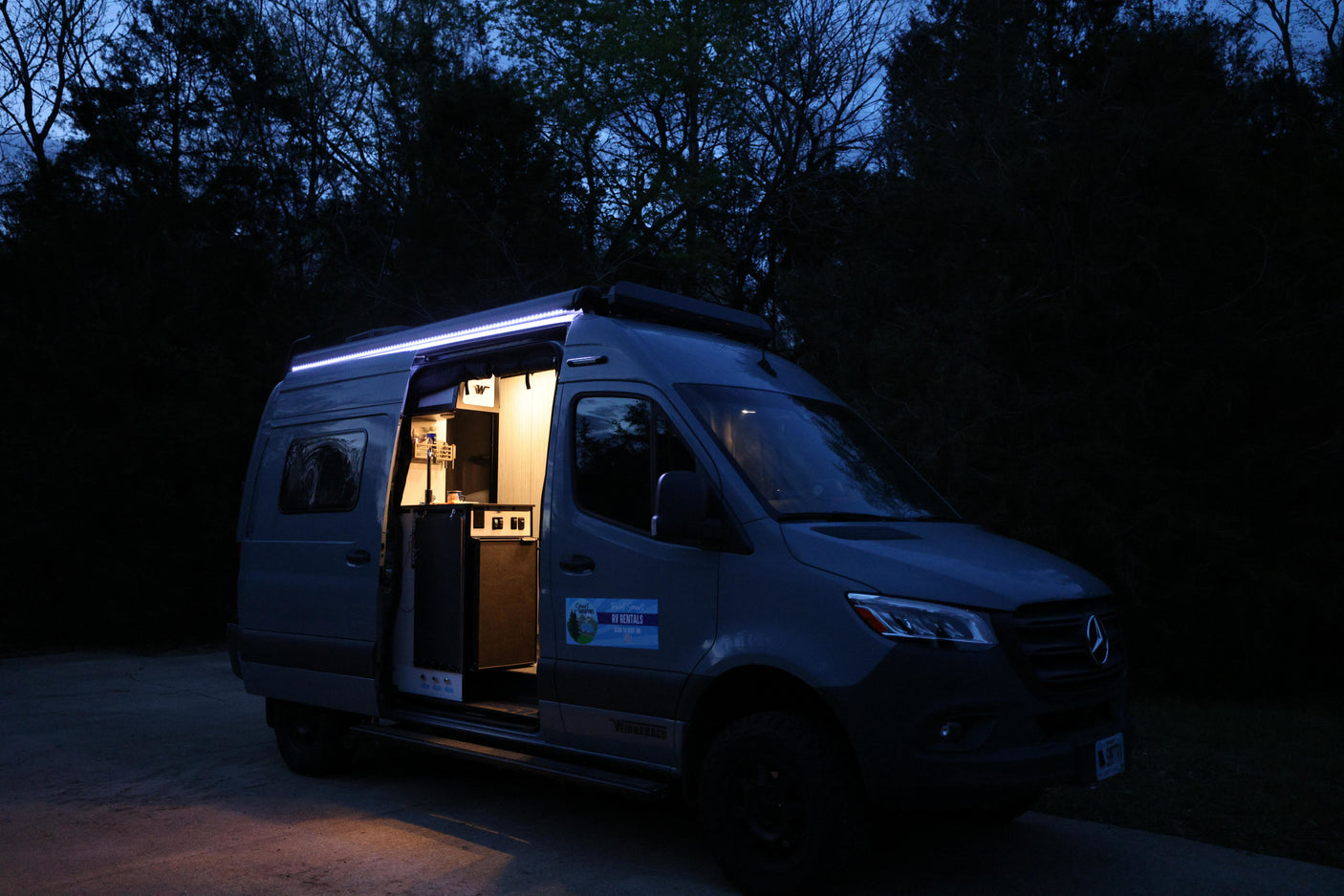 Grey Mercedes Sprinter RV rental parked at night, side door open with lit interior & bright exterior LED strip.