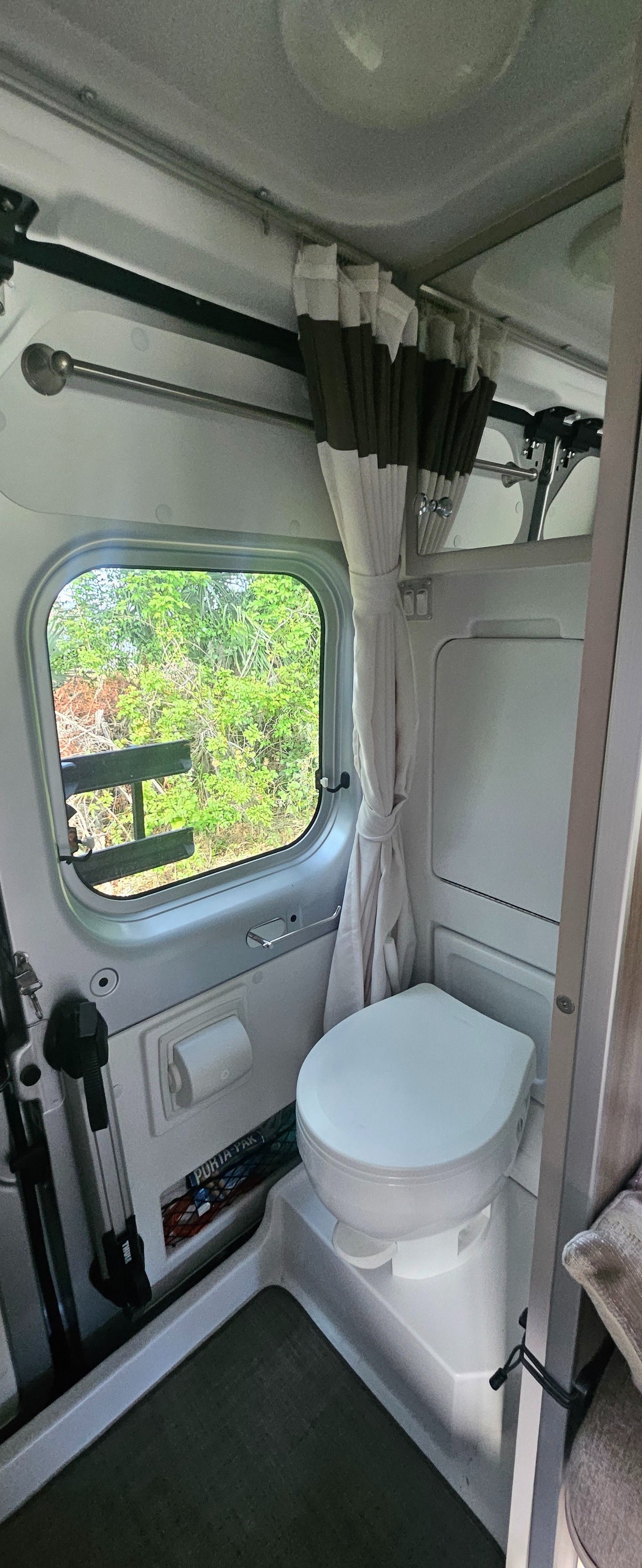 Compact RV rental wet bath: white toilet, tied-back shower curtain, and a window framing lush green nature outside.