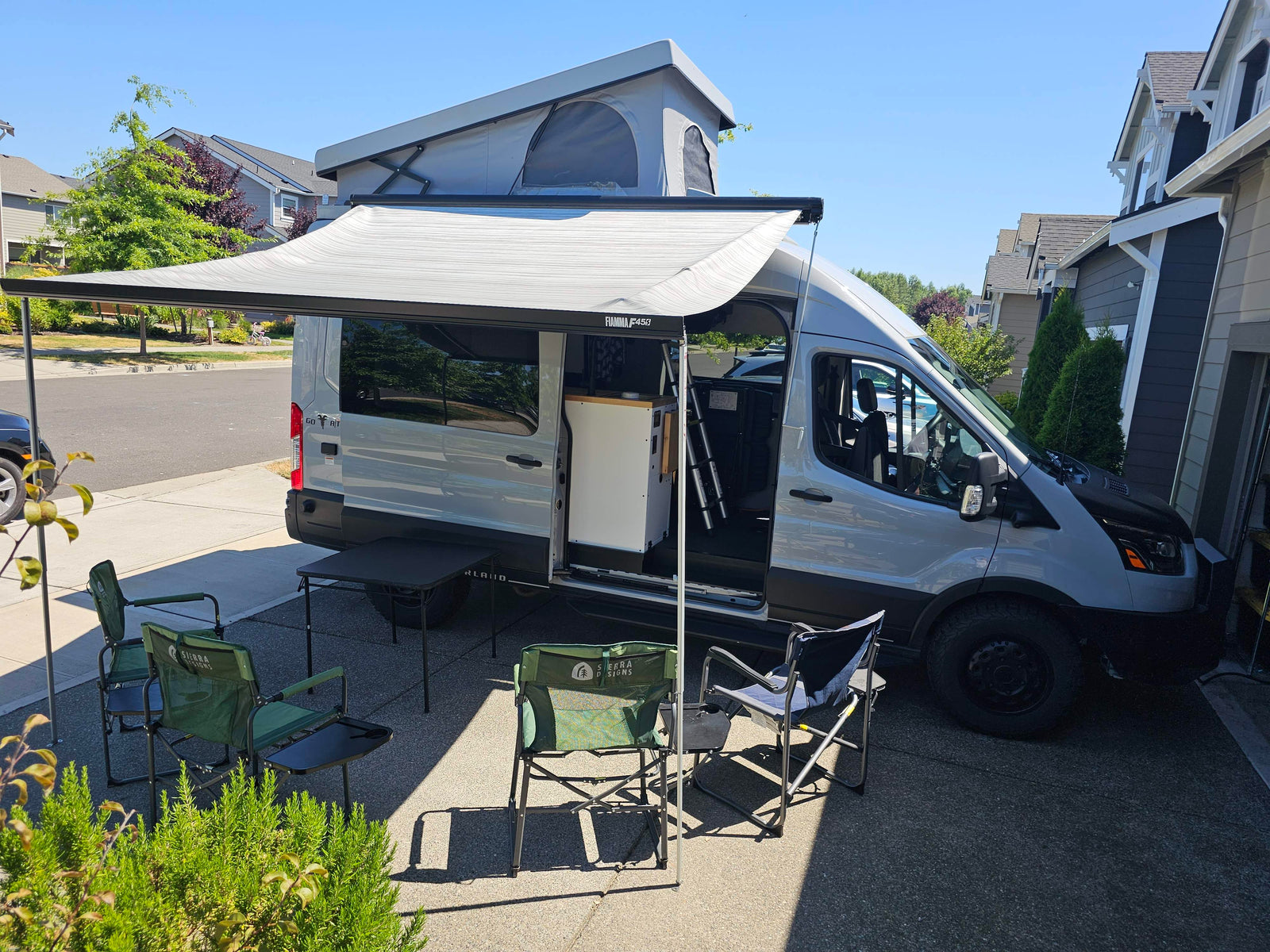 Gray RV rental campervan with pop-top roof and extended awning, set up with camping chairs and table for outdoor comfort.
