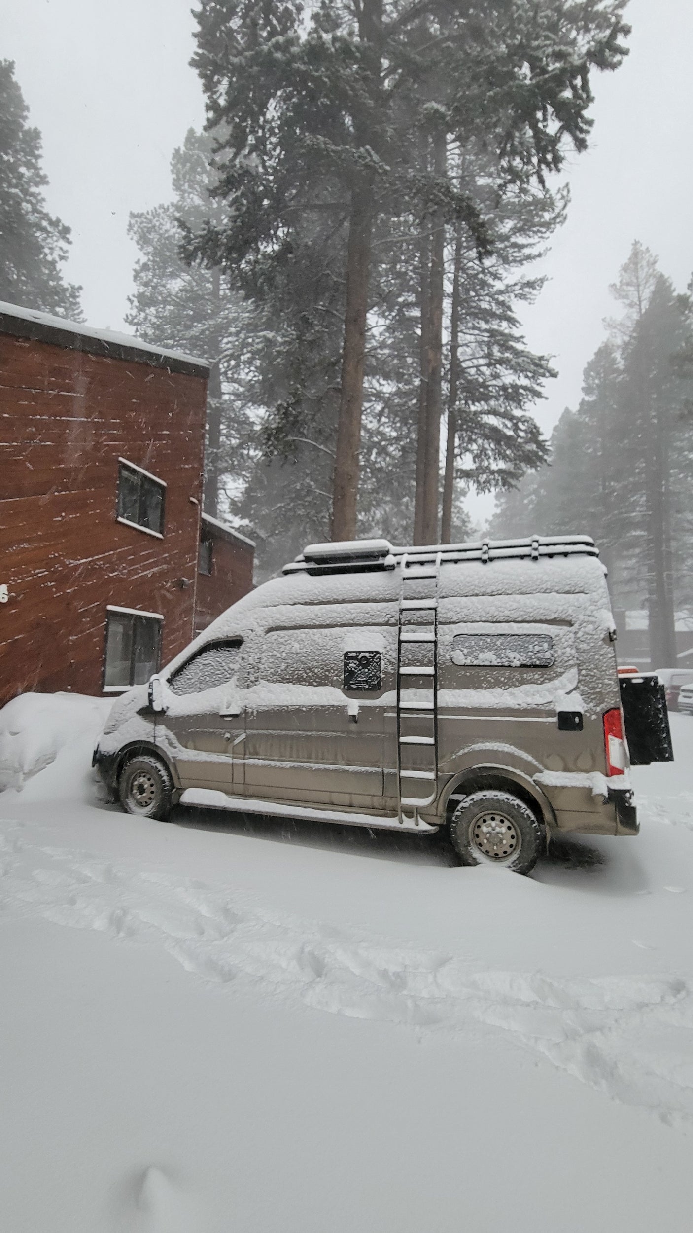 A snow-covered RV rental van is partially buried in deep snow, parked next to a building and tall trees.