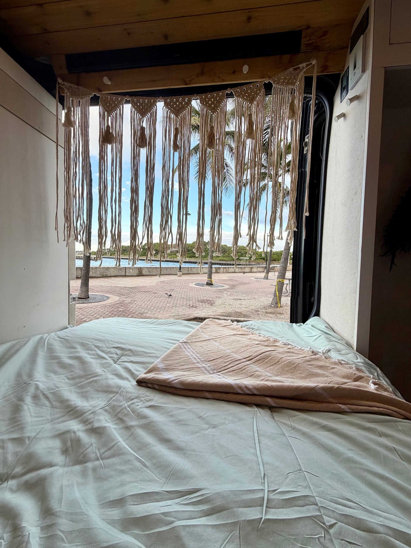 Cozy RV rental bed with a macrame curtain, offering a tropical waterfront view with palm trees. Ideal for van life.