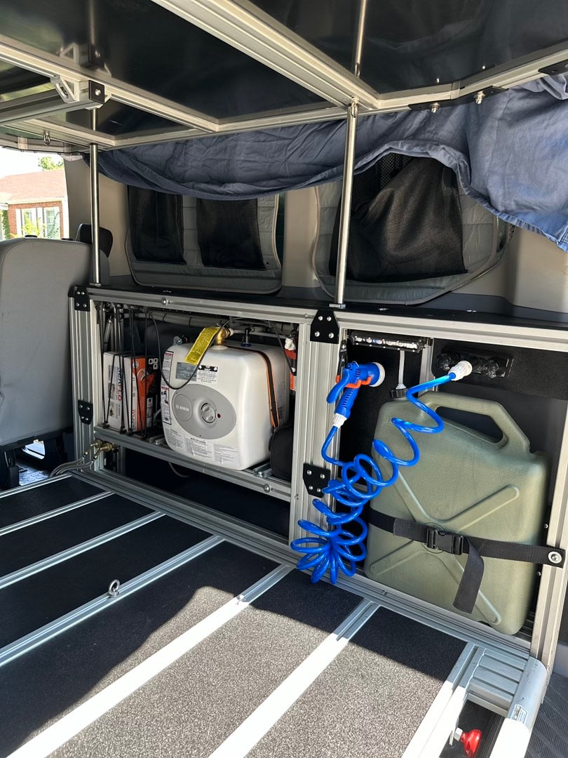 An RV rental's compact utility area shows a Bosch water heater, a green water jug with a blue shower hose, and aluminum shelving.